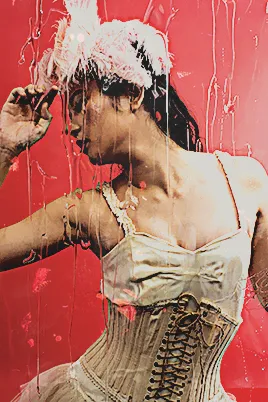 tamura meimi posing in a ballerina-like outfit with a semi-elaborate feathery headpiece, against a red background. there is a rainwater overlay from the original photo. the image is edited to add various effects and a grunge-y flower petal texture
